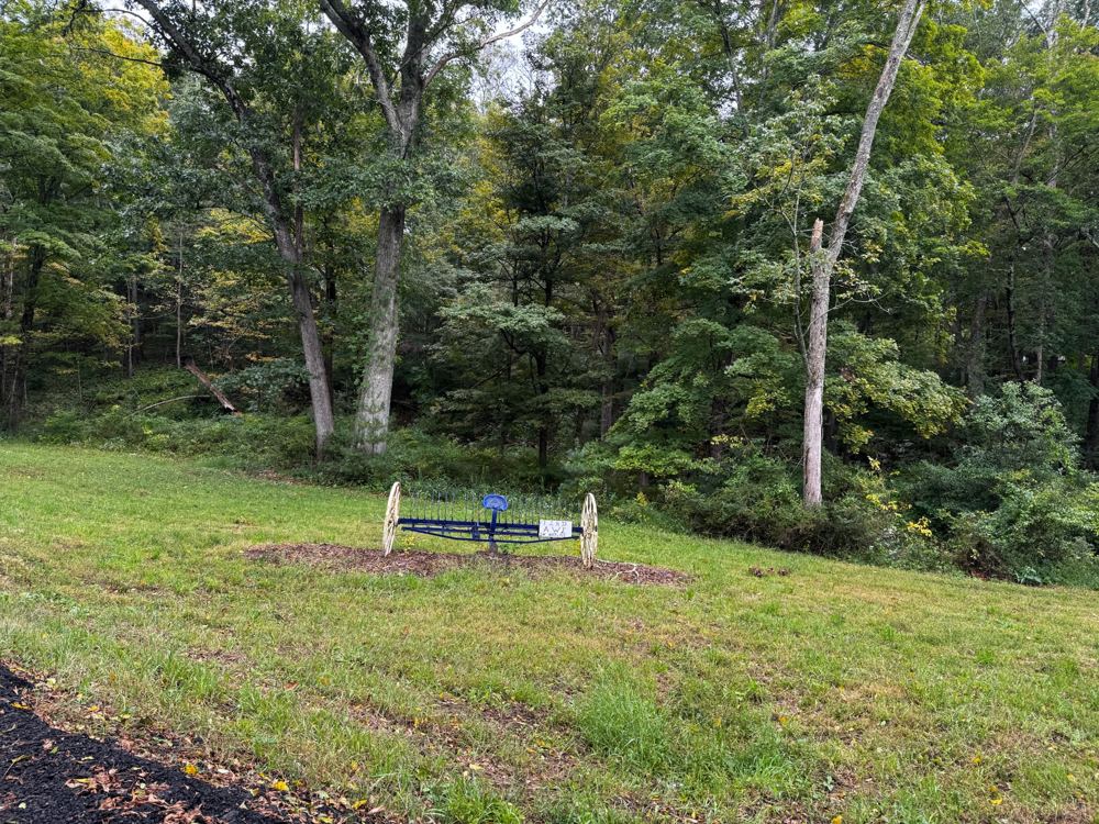 An antique blue hay rake with cream-colored spoked wheels stands on a mulched patch of lawn in front of a wooded hillside, with a small sign reading Find awe in nature